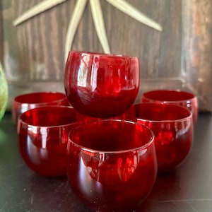 May include: A collection of six small, rounded drinking glasses made of translucent red glass. The glasses feature a smooth, curved shape with a wide opening and a solid base, arranged in a cluster to showcase their vibrant, deep red color.