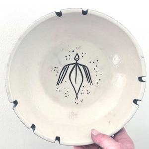 May include: Off-white ceramic bowl with a black abstract figure design in the center, surrounded by small black dots. The rim has a dark blue trim and several cut-out notches. The bowl is held by a person's hand.