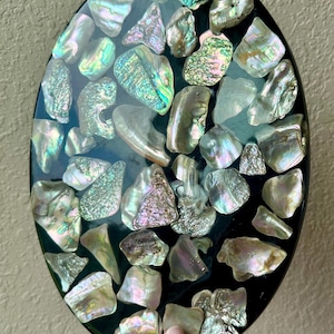 May include: Oval-shaped decorative piece with a black background, inlaid with iridescent abalone shells. The shells vary in size and shape, displaying a range of colors including green, blue, and purple. The piece is held by a hand.