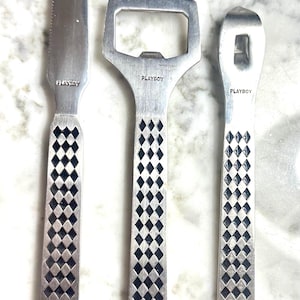 May include: Set of three stainless steel bar tools, including a cheese knife, bottle opener, and can opener. Each tool features a diamond pattern on the handle and the "PLAYBOY" logo.