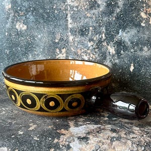 May include: A ceramic bowl with a handle, featuring a brown interior and a black and yellow circular pattern around the rim. The handle is dark brown, and the bowl has a glossy finish. The bowl is likely for serving or display.