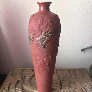May include: A tall, slender terracotta-colored vase with a textured, matte surface. The body of the vase features a raised, metallic-toned relief of a dragon with detailed scales and claws wrapping around the center of the cylindrical vessel.