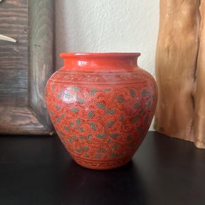 May include: A round, bulbous vase with a vibrant orange base color. The surface is decorated with an intricate, repeating pattern of etched green and gold-toned leaves and swirling vines that cover the entire exterior of the vessel.