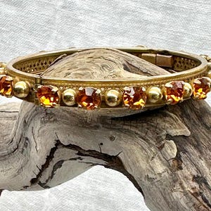 May include: A gold-toned bangle bracelet with a decorative design and a row of amber-colored rhinestones.