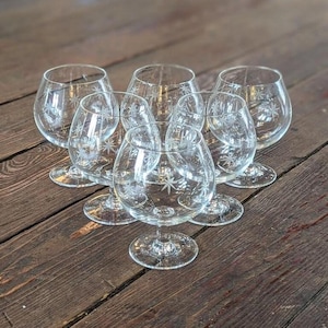May include: Six clear brandy snifters with etched starburst designs. The glasses are arranged on a dark wooden surface, showcasing their elegant shape and intricate detailing. The stemware is ideal for serving spirits.
