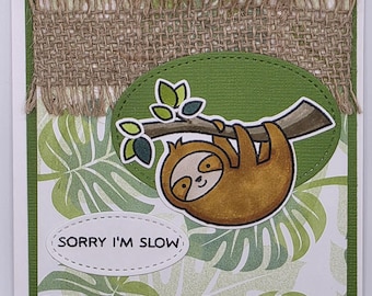 May include: A handmade greeting card featuring a cartoon sloth hanging from a branch. The sloth is brown with a smiling face. The card has a green border, a burlap accent, and a background of green leaves. The text "SORRY I'M SLOW" is on the card.