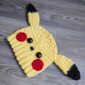 May include: A yellow crocheted hat with black ears and red cheeks. The hat is shaped like a Pikachu, a popular cartoon character.