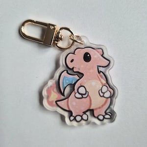May include: A pink cartoon dragon keychain with a gold-colored clasp. The dragon has a light pink body, a yellow belly, and a blue and yellow wing. The keychain is made of clear acrylic.