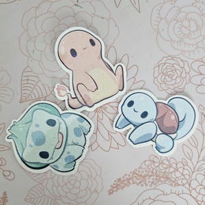 May include: Three cartoon character stickers featuring a light blue turtle, a light green frog, and a light orange salamander. The stickers are on a light pink background with a floral pattern.