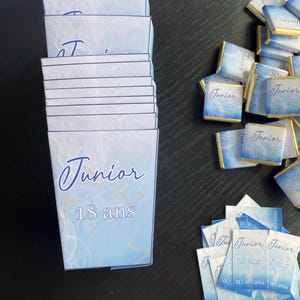 Peut inclure: Une pile de boîtes de faveur bleues et blanches avec le texte "Junior 18 ans" sur le devant. Il y a aussi de petites barres de chocolat bleues et blanches avec le texte "Junior 18 ans" dessus. Il y a aussi de petites cartes bleues et blanches avec le texte "Junior 18 ans" et la date "12/10/2024" dessus.