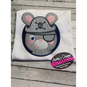 Pode incluir: Uma camiseta branca com um design bordado de um rato cinza usando um chapéu de pirata xadrez azul e branco com uma caveira e ossos cruzados. O rato tem um tapa-olho rosa e orelhas rosas.