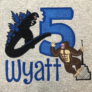 Può includere: Una felpa grigia con testo ricamato in blu che dice "Wyatt" e un numero blu "5". Un mostro nero simile a Godzilla è ricamato sul lato sinistro del numero e un mostro marrone simile a King Kong è ricamato sul lato destro del numero.