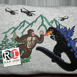 Puede incluir: Sudadera gris bordada con un monstruo Godzilla negro y azul con una llama roja, luchando contra un monstruo King Kong marrón en un paisaje montañoso. La sudadera también presenta un logotipo para "RAN-DOM DESIGNS" en rojo, azul y blanco.