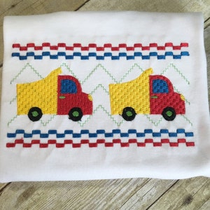 Pode incluir: Camiseta branca bordada com uma borda vermelha, branca e azul e dois caminhões basculantes amarelos, vermelhos e azuis.
