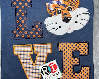 May include: A navy blue sweatshirt with the word "LOVE" in orange and blue patterned letters. The "O" is replaced with a cartoon tiger face with an orange and blue bow.