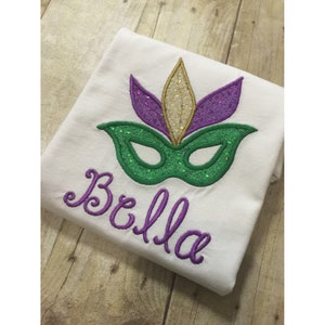 May include: A white t-shirt with a purple and green Mardi Gras mask and the name "Bella" embroidered in purple.