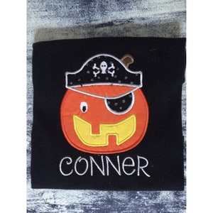 May include: Black t-shirt featuring an orange pumpkin wearing a pirate hat and an eye patch. The pumpkin has a yellow smile and the text "CONNER" is below the pumpkin.