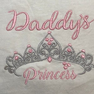 May include: Embroidered design on a white fabric with the text "Daddy's Princess" in pink thread. A silver crown with pink accents is centred below the text.