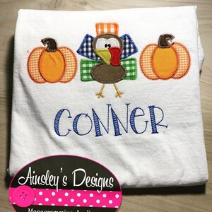 Puede incluir: Camiseta blanca con una aplicación de pavo marrón, dos calabazas naranjas y el nombre "CONNER" bordado en hilo azul. La camiseta también tiene una etiqueta de lunares rosa y blancos con el texto "Ainsley's Designs".