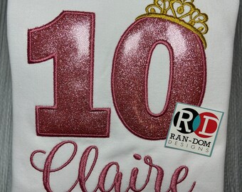 Peut inclure: Sweat-shirt blanc avec le chiffre "10" rose scintillant, surmonté d'une couronne dorée. Le nom "Claire" est brodé en rose en dessous. Une étiquette avec "RAN-DOM DESIGNS" est dans le coin inférieur droit.
