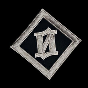 Puede incluir: Parche negro con borde de diamante bordado en plata. Dentro del diamante hay un cuadrado negro con una letra "M" estilizada bordada en plata. El parche está sobre un fondo negro.