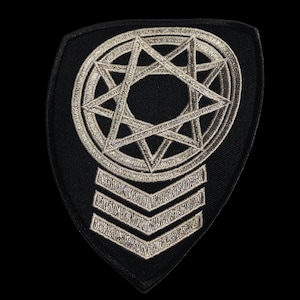 Puede incluir: Parche bordado negro con un diseño de estrella de siete puntas plateada dentro de un círculo, sobre tres rayas en forma de cheurón. El parche tiene forma de escudo y es para ropa o accesorios.