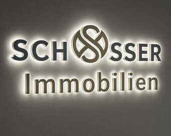 Pode incluir: Letreiro iluminado com o nome "SCHOSSER" acima da palavra "Immobilien". As letras são em cinza escuro e castanho, com contorno branco brilhante. O logotipo é um "S" estilizado entrelaçado com um círculo.