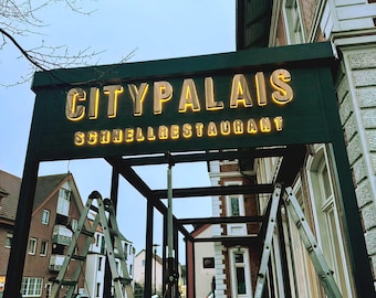 Pode incluir: Letreiro de restaurante "CITYPALAIS SCHNELLRESTAURANT" com letras douradas iluminadas. O letreiro está montado numa estrutura verde escura. Escadas e andaimes em primeiro plano sugerem obras ou manutenção em curso.