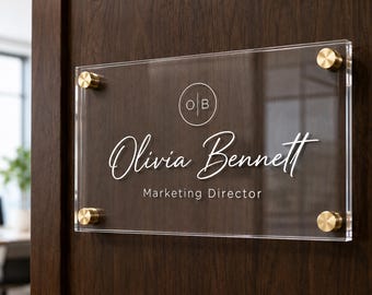 Pode incluir: Placa de escritório em acrílico transparente com espaçadores dourados. A placa exibe o nome "Olivia Bennett" em fonte cursiva, o título "Marketing Director" e as iniciais "OB" em um círculo.
