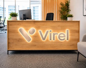 Pode incluir: Uma receção de madeira com a palavra "Virel" iluminada em letras prateadas. A secretária tem um acabamento em grão de madeira clara e está situada num espaço de escritório moderno com plantas e um monitor de computador.