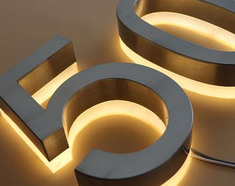 Pode incluir: Close-up dos números metálicos iluminados "50". Os números têm um acabamento em metal escovado com um brilho amarelo quente que emana de dentro. O design é moderno e elegante, adequado para decoração de casa ou escritório.