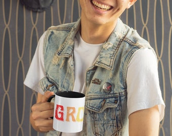 May include: A white ceramic mug with a black interior, featuring the word "GRO" in red, orange, and yellow lettering. The mug is held by a person wearing a denim vest and a white t-shirt. The background includes a decorative wall.