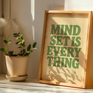 Può includere: Una stampa incorniciata con le parole "MIND SET IS EVERYTHING" in lettere verdi su sfondo crema. Una pianta in vaso con foglie verdi si trova a sinistra della cornice. La cornice è in legno chiaro.