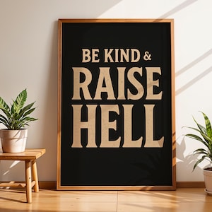 May include: A framed art print with the words "BE KIND & RAISE HELL" in a bold, serif font against a black background. The frame is a warm wood tone. Two potted plants flank the artwork, one on a small wooden stool.