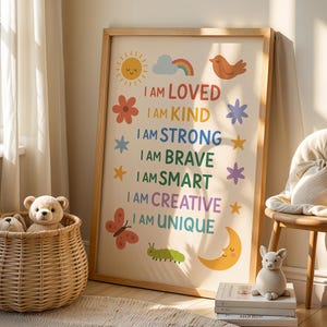 May include: A framed print with the words "I AM LOVED, I AM KIND, I AM STRONG, I AM BRAVE, I AM SMART, I AM CREATIVE, I AM UNIQUE" in colourful text. The print features illustrations of a sun, rainbow, bird, flower, stars, butterfly, caterpillar, and moon. A wicker basket with stuffed animals is in the foreground.