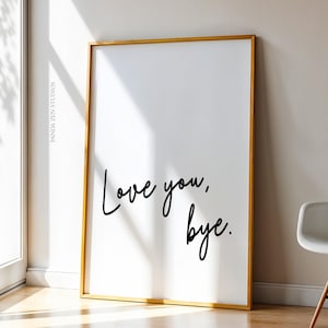 Op de afbeelding: Een ingelijste kunstdruk met een witte achtergrond en een gouden lijst. De zwarte cursieve tekst luidt "Love you, bye." De print leunt tegen een muur naast een raam en een witte stoel.