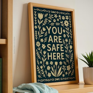 May include: A framed print with the text "YOU ARE SAFE HERE" in cream, surrounded by floral designs on a dark teal backdrop. The frame is light wood. A small potted plant is also visible. The print is on a shelf.