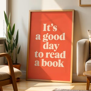 Puede incluir: Un póster enmarcado con el texto "It's a good day to read a book" en blanco sobre un fondo coral. El póster está apoyado contra una pared en una habitación con una silla de madera, plantas y un sofá.