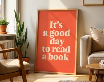 May include: A framed poster with the text "It's a good day to read a book" in white on a coral background. The poster is leaning against a wall in a room with a wooden chair, plants, and a sofa.