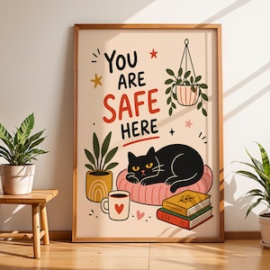 May include: A framed wall art print with a cream background featuring the black text "YOU ARE SAFE HERE" in a bold font. The illustration includes a black cat resting on a pink cushion, a hanging plant, a potted plant, a coffee mug, and two books.