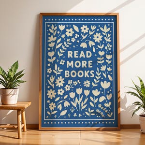 Könnte beinhalten: Ein gerahmter Kunstdruck mit blauem Hintergrund und floralem Design. Die Worte "READ MORE BOOKS" stehen im Zentrum. Der Rahmen ist braun. Zwei Topfpflanzen und ein kleiner Holztisch sind im Bild.