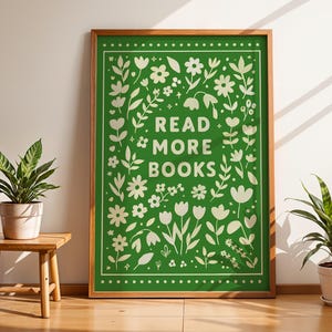 May include: A framed green poster with the words "READ MORE BOOKS" in white, surrounded by floral and leaf illustrations. The poster is in a wooden frame and is displayed against a white wall.