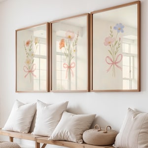Puede incluir: Tres impresiones artísticas florales enmarcadas. Cada impresión presenta un ramo de flores en acuarela atado con una cinta rosa. Los marcos son de color marrón cálido. Las impresiones se exhiben sobre un banco de madera con almohadas.