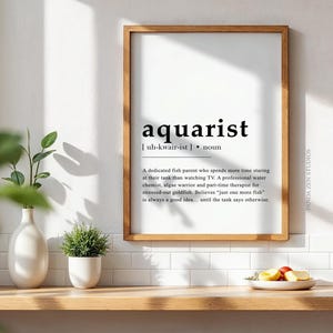 Op de afbeelding: Zwart-witte printbare kunstprint met de definitie van het woord "aquarianer" in een minimalistische stijl. De definitie luidt: "Een toegewijde visouder die meer tijd besteedt aan het kijken naar zijn aquarium dan aan het kijken naar tv. Een professionele waterchemicus, algenstrijder en parttime therapeut voor gestreste goudvissen. Gelooft dat "nog maar één vis" altijd een goed idee is... totdat de tank anders zegt."
