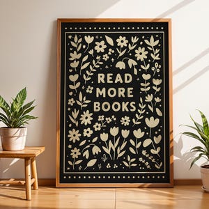 Könnte beinhalten: Ein gerahmter schwarz-goldener Druck mit den Worten „READ MORE BOOKS“, umgeben von floralen und Blattillustrationen. Der Druck wird an einer Wand neben einem kleinen Holztisch mit einer Topfpflanze und einer weiteren Topfpflanze auf dem Boden angezeigt.