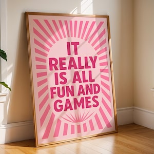 May include: A framed pink art print with the words "IT REALLY IS ALL FUN AND GAMES" in a bold, playful font. The design features a sunburst pattern in varying shades of pink. The artwork is displayed on a wooden floor.