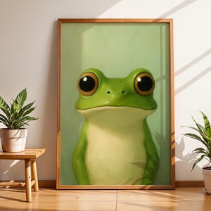 May include: A framed art print featuring a large, cartoon-style frog with big, expressive eyes. The frog is primarily green with a lighter belly, set against a light green background. The artwork is displayed in a room with plants and a wooden side table.