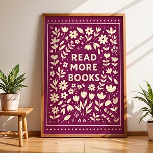May include: A framed art print with the words "READ MORE BOOKS" in white, surrounded by floral and leaf designs on a deep purple background. The print is in a wooden frame and is displayed next to a small wooden table with a potted plant.