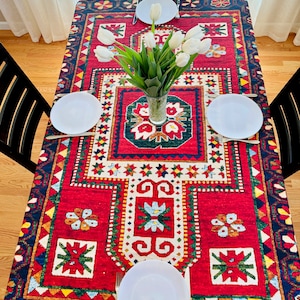 May include: A rectangular red tablecloth with a detailed geometric pattern in blue, white, green, and yellow. The table is set with white plates, silverware, and a vase of white tulips. Black chairs surround the table.