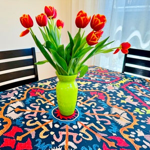 Könnte beinhalten: Ein Strauß rot-gelber Tulpen in einer hellgrünen Vase auf einem Tisch. Der Tisch ist mit einer dunkelblauen, gemusterten Tischdecke bedeckt, und im Hintergrund sind zwei schwarze Stühle zu sehen. Die Szene wird von natürlichem Licht beleuchtet.